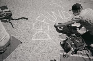 A photograph of a person kneeling on a blanket laid out on pavement and writing &quot;DEFUND&qu ...