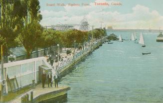 Picture of people walking on a boardwalk with the lake and sail boats to the right and trees on ...
