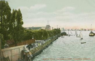 Picture of people walking on a boardwalk with the lake and sailboats on the right and trees on …