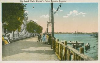 Picture of a wide boardwalk and people sitting and walking next to a lake with boats. 