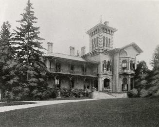 A photograph of a large two-story house, with third floor consisting of a tower. There are wide ...