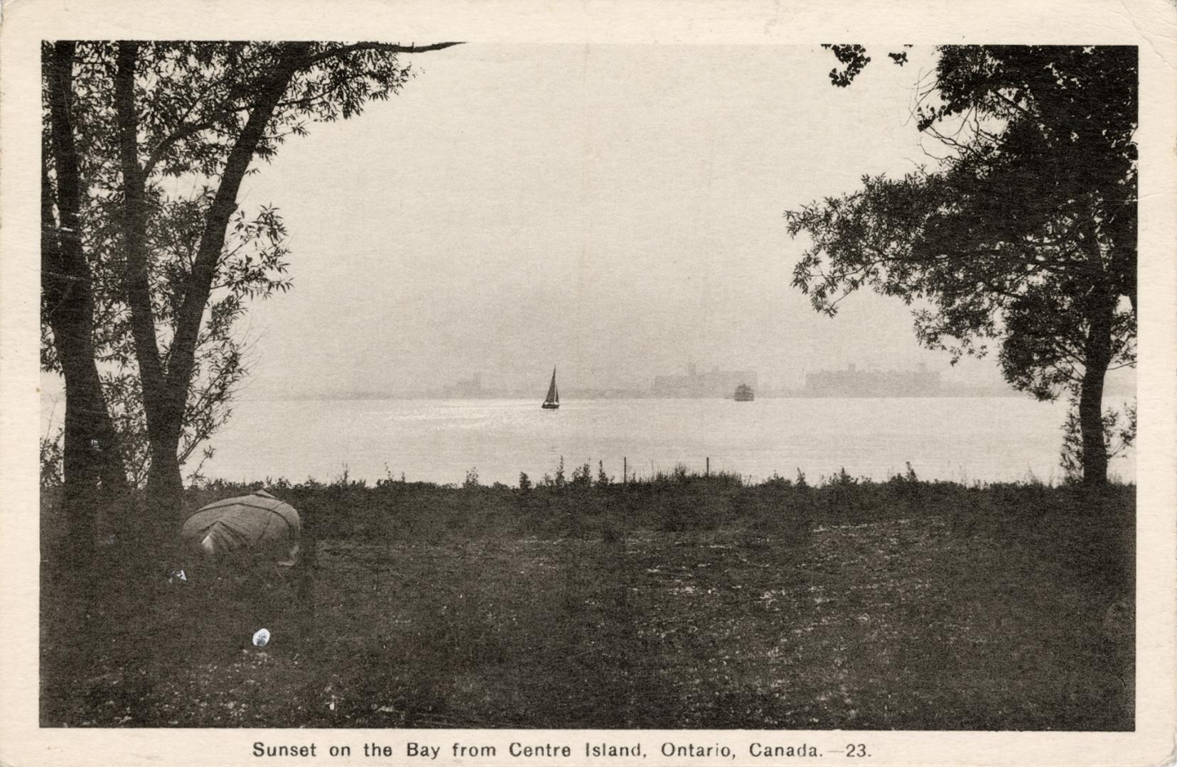 Hazy view of Toronto skyline with sailboat in the distance and canoe on grass in foreground. 