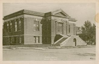 Picture of two storey public library with two sided front steps and house and tree on right. 