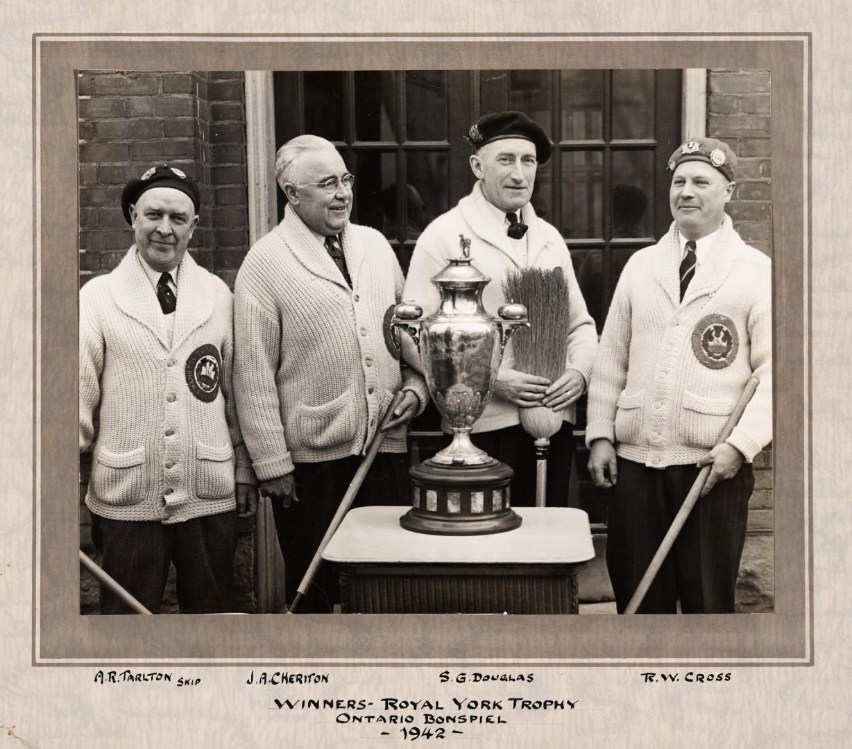 A photograph of four men holding curling brooms standing in front of a pedestal with a trophy r…