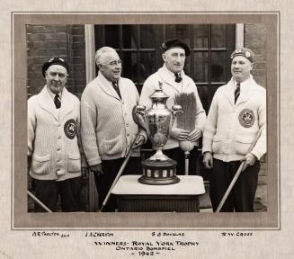A photograph of four men holding curling brooms standing in front of a pedestal with a trophy r ...