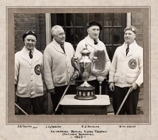 A photograph of four men holding curling brooms standing in front of a pedestal with a trophy r…