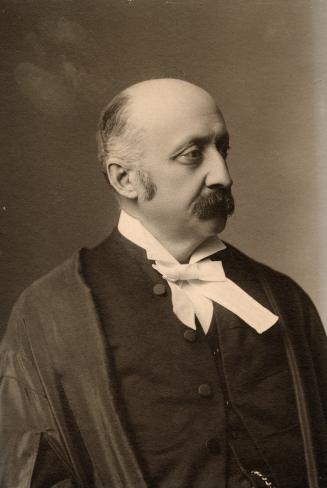 A formal photograph of a middle-aged man, taken in a studio. The man is balding, has a moustach…