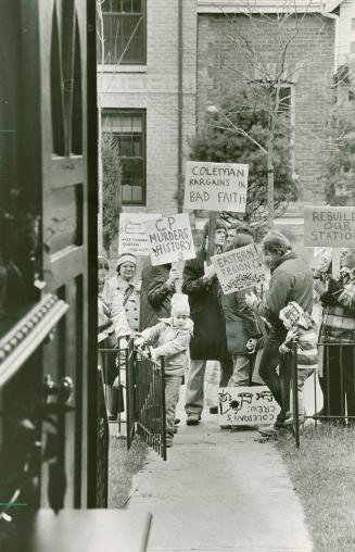 A photograph of a group of about twelve protestors standing on a sidewalk in front of a gate. T ...