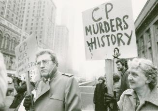 A photograph of a group of people holding picket signs in front of a building. On the left a mi ...