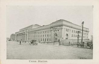 A photograph of a railway station, with a wide city street in front of it. There are cars, a ho ...