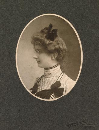 A photograph of a young woman, taken at a photography studio. She is wearing a dress with a hig…