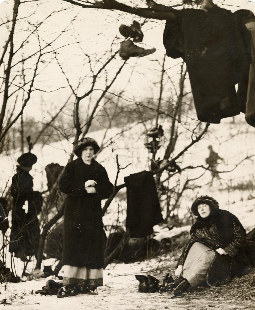 A photograph of two women, one sitting and the other standing underneath a tree in winter. Ther…