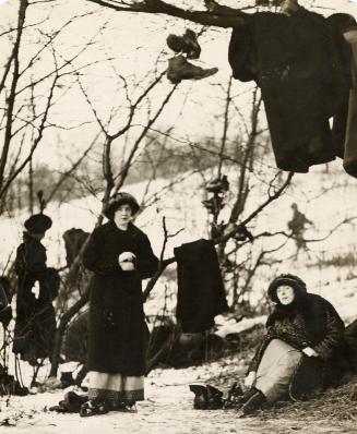A photograph of two women, one sitting and the other standing underneath a tree in winter. Ther…