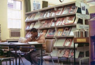 Picture of man reading at a table in a library with shelves of magazines behind him. 