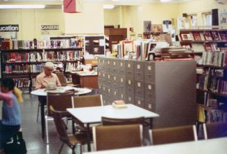 Picture of a man sitting at a round table in a crowded library reading room with filing cabinet ...