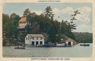 Several buildings and boathouses on a rocky ledge in front of a body of water with people in bo…