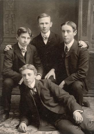 A photograph of three teenage boys and one male adult, wearing formal suits and ties. Two of th…