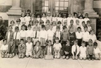 A class photograph of a group of children posing together on the front steps of the school. The ...