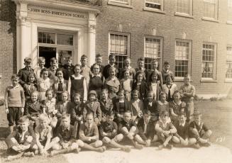 A class photograph of a group of students sitting and standing in front of the front door of th ...