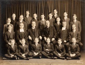 A formal class photograph, taken in a studio, of twenty-four students and teachers. The student ...