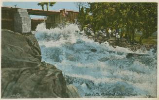 Colorized photograph of a bridge spanning a waterfall. Rocks in the foreground.