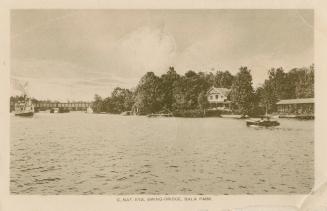 Black and white photograph of a motor boat on a lake with cottages on the shoreline. Bridge in  ...