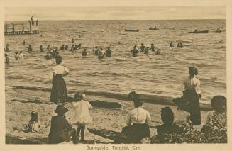 Women and children on a beach watch people swimming in a lake. 