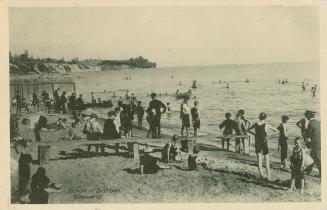 A crowd of people stand and sit on a beach with others swimming in a lake. 