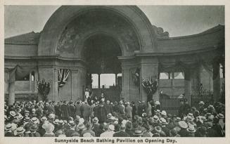 A huge crowd of people and dignitaries stand in front of a very large stone arch.