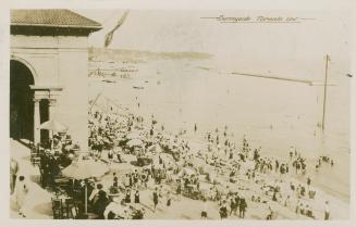 Crowds of people on a beach at a lake with a building in the left foreground. 