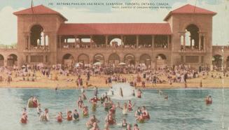 Picture of people swimming in lake at a beach with a large bathing pavilion behind. 