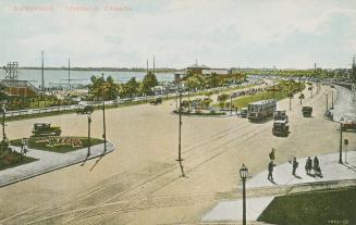 Picture of a long roadway with some cars and a streetcar running beside a lake. 