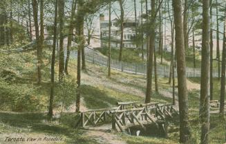 Picture of a log bridge in a wooded area with large houses in the background.
