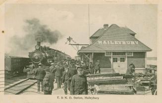 Black and white photograph of a train on the tracks beside a station with many people on the pl ...