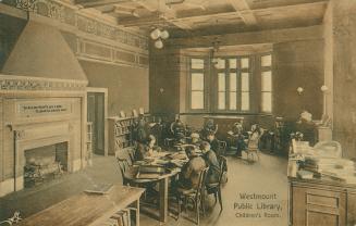 Picture of children seated reading at a large table next to a big fireplace in a children's lib ...