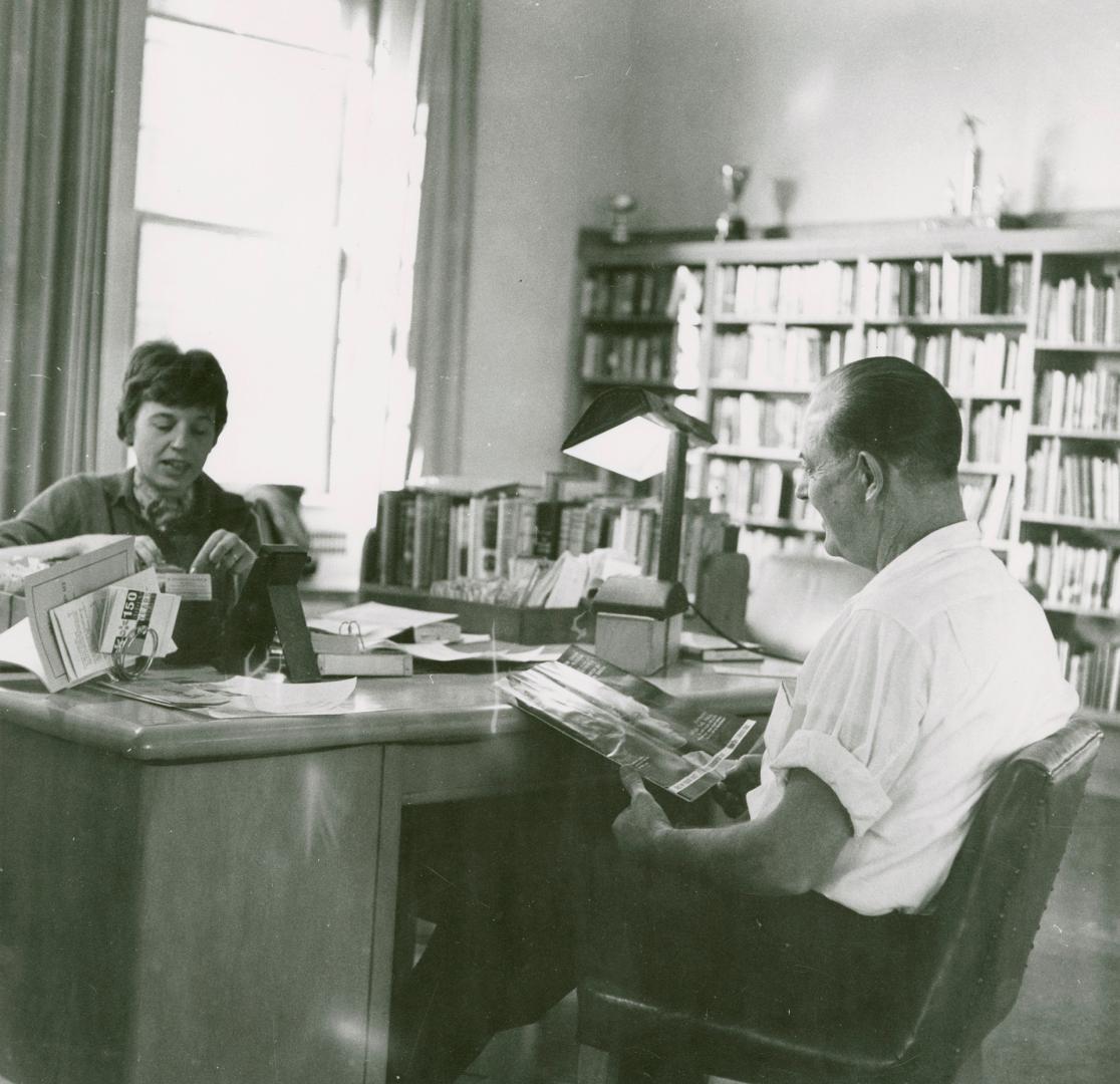 A librarian sits at a desk speaking with a man seated in front of her. 