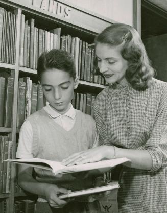 A woman shows an open book to a boy with bookshelves in background. 