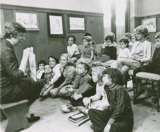 A librarian reads a story book to a group of children seated in front of her on the floor and o ...