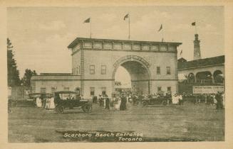 Picture of arched entrance gate with flags on top and cars and people in front. 