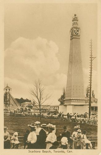 People standing in front of a tall stone monument with amusement park buildings in background. 