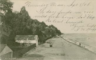 Black and white photograph of a long stretch of beach with a fence and buildings to the left.