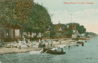 People and boats along a sandy shoreline with building to the left of the picture.