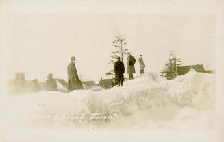ix people standing on a bank of snow and ice on a beach with houses in the background.