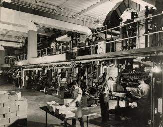 A photograph of a a large printing press, with several people working at and around it. In the  ...