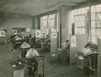 A photograph of an office with warehouse-style windows on the right side and men and women work ...