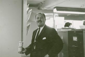 A photograph of a man wearing a business suit and tie standing in an office in front of some fi ...