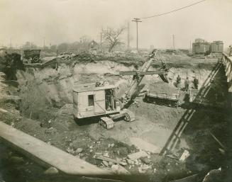 A photograph of a construction site, with a backhoe piling dirt into a flatbed truck inside a p ...