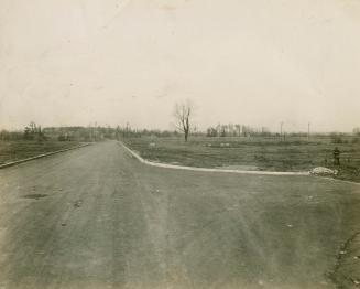 A photograph of a semi-rural area, with a curbed dirt road, electrical poles and wires, and a f ...