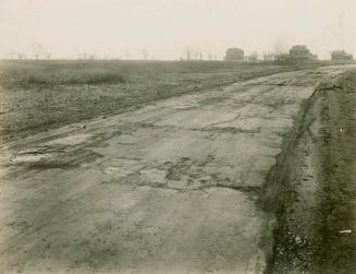 A photo of a cracked asphalt road running through the middle of a field, with three large house ...