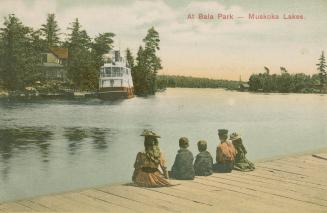 Five children are sitting on a wooden dock watching a steamship on the other side of a lake.